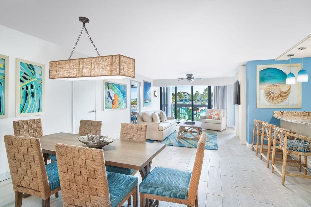 A modern living room with a dining area, featuring wicker furniture, a large window with a view of a pool, and artwork on the walls.