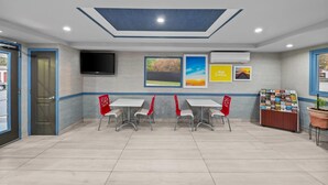 A modern indoor space with a flat ceiling, recessed lighting, and a wall-mounted TV. There are three tables with red chairs, a bulletin board with various items, and a potted plant. - Days Inn by Wyndham Chesapeake (Chesapeake)