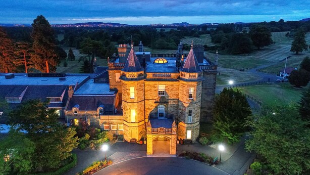 A large, historic building with multiple towers and a central courtyard, surrounded by trees and a well-maintained lawn.