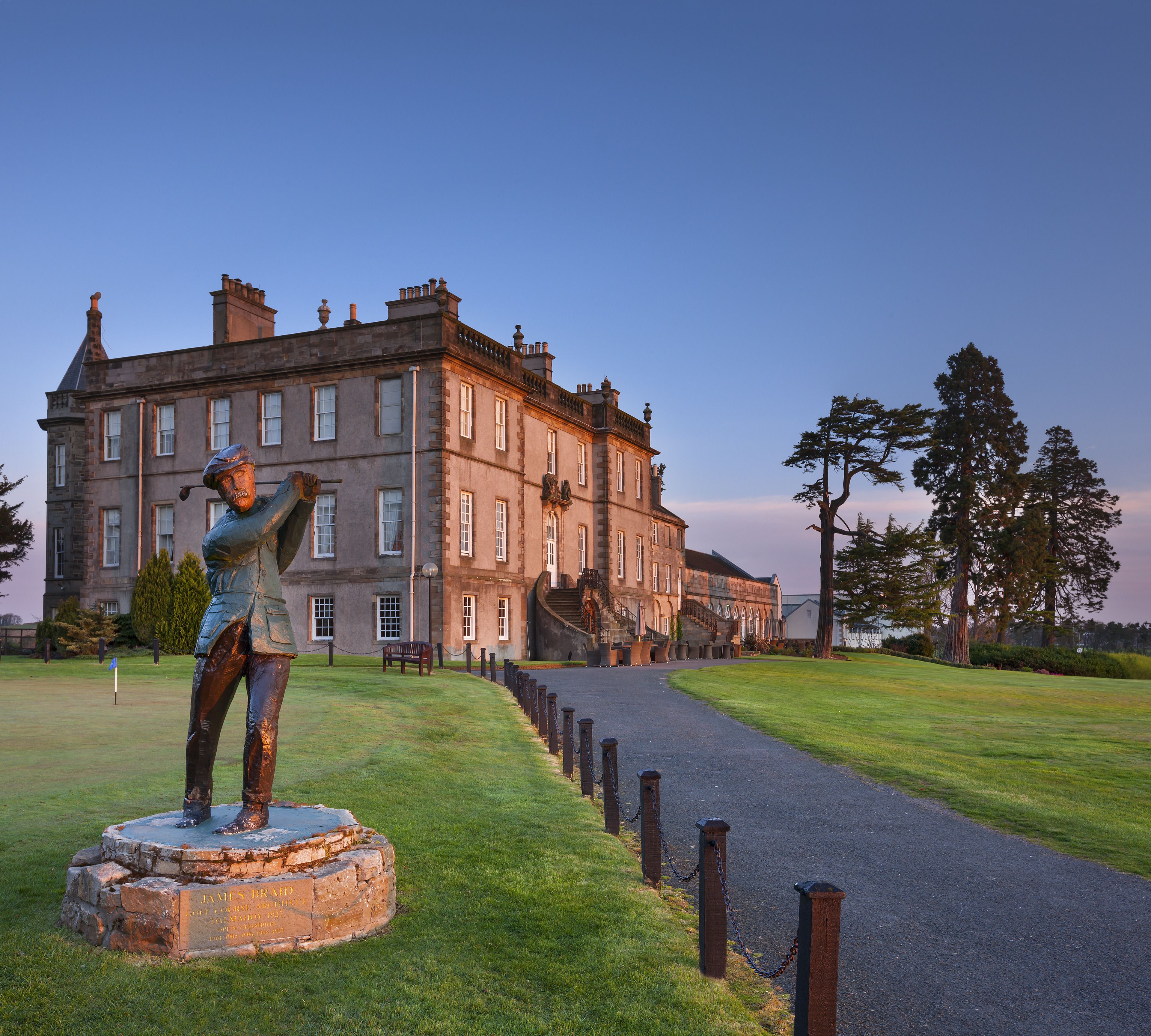 A statue of a golfer on a pedestal in front of a large, historic building with a paved path leading to it.