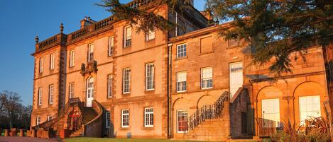 A large, historic building with multiple windows, a central staircase, and a well-maintained lawn.