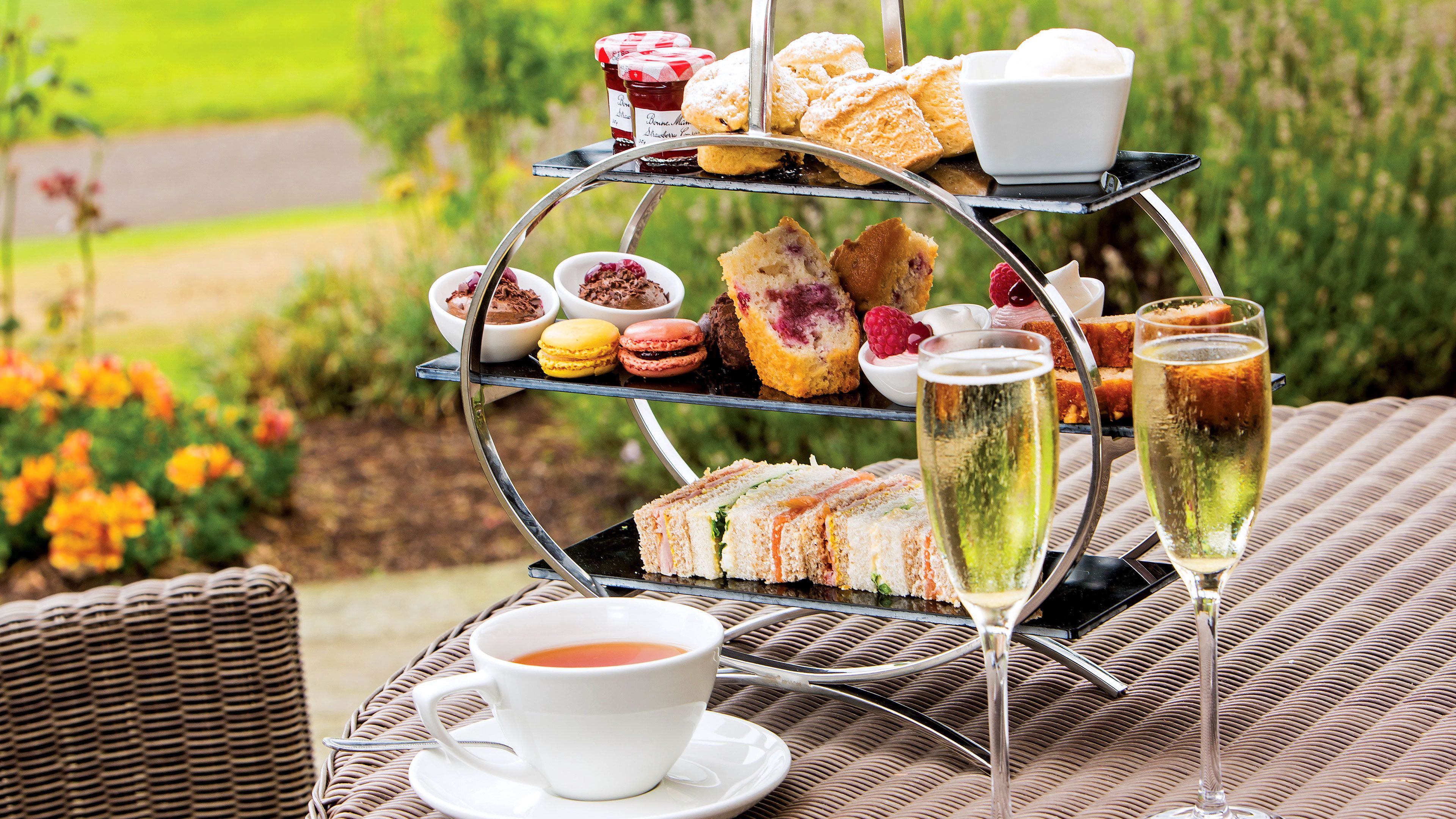 A three-tiered outdoor tea and snack stand with sandwiches, pastries, and desserts, accompanied by two glasses of champagne and a cup of tea.