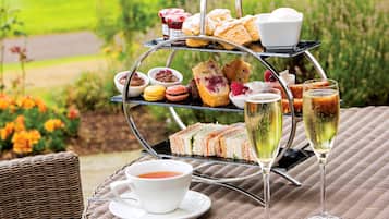 A three-tiered outdoor tea and snack stand with sandwiches, pastries, and desserts, accompanied by two glasses of champagne and a cup of tea.