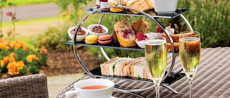 A three-tiered outdoor tea and snack stand with sandwiches, pastries, and desserts, accompanied by two glasses of champagne and a cup of tea.