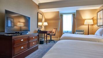 A hotel room with a flat-screen TV, a desk with a chair, two beds, and a large window with curtains.