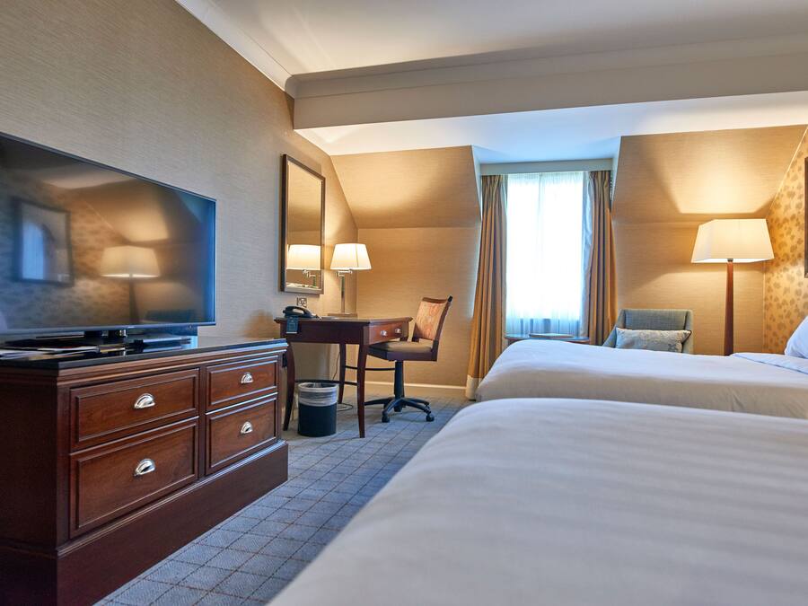 A hotel room with a flat-screen TV, a desk with a chair, two beds, and a large window with curtains.