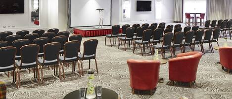 A conference room with rows of chairs, a stage with a red backdrop, and a large screen.