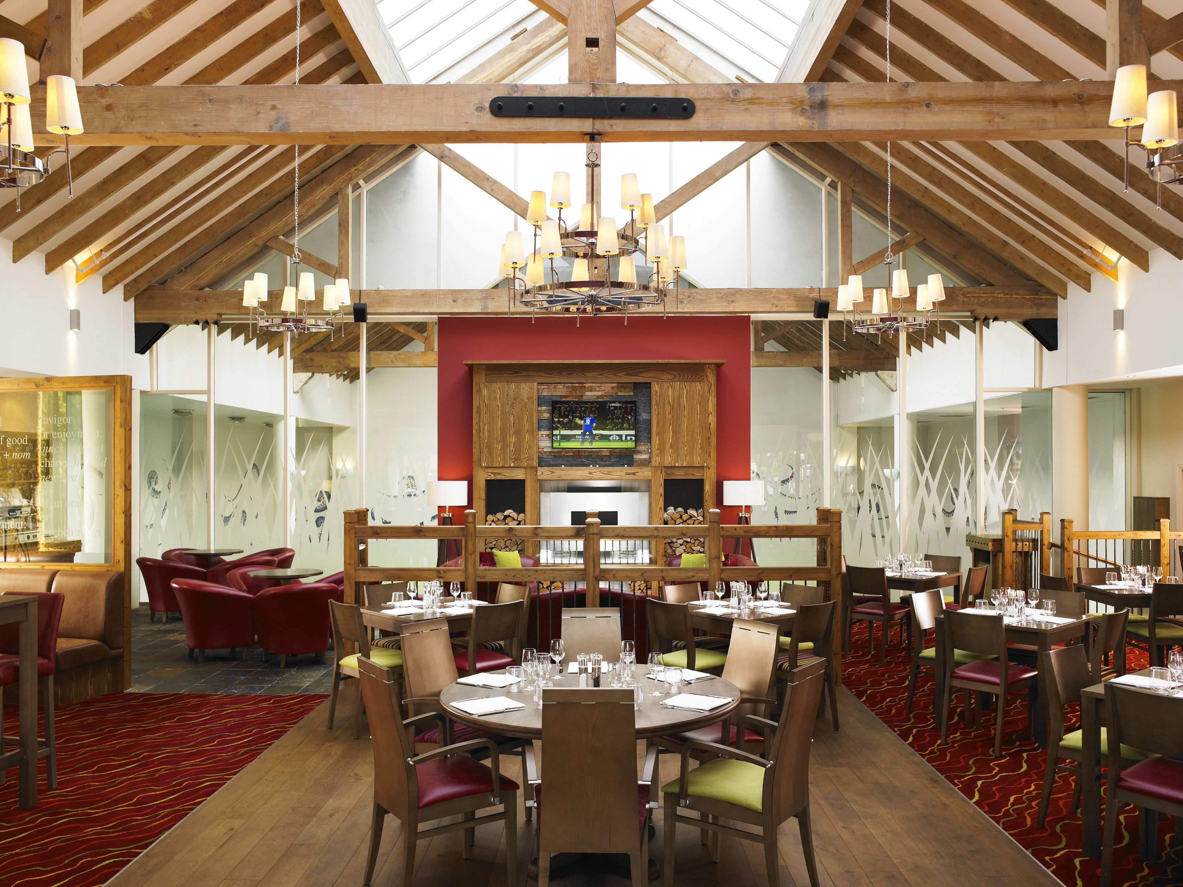 Un restaurant avec des poutres en bois, des chaises à l’assise rembourrée de tissu rouge et une cheminée centrale. Les tables sont dressées avec des verres à vin et des menus.