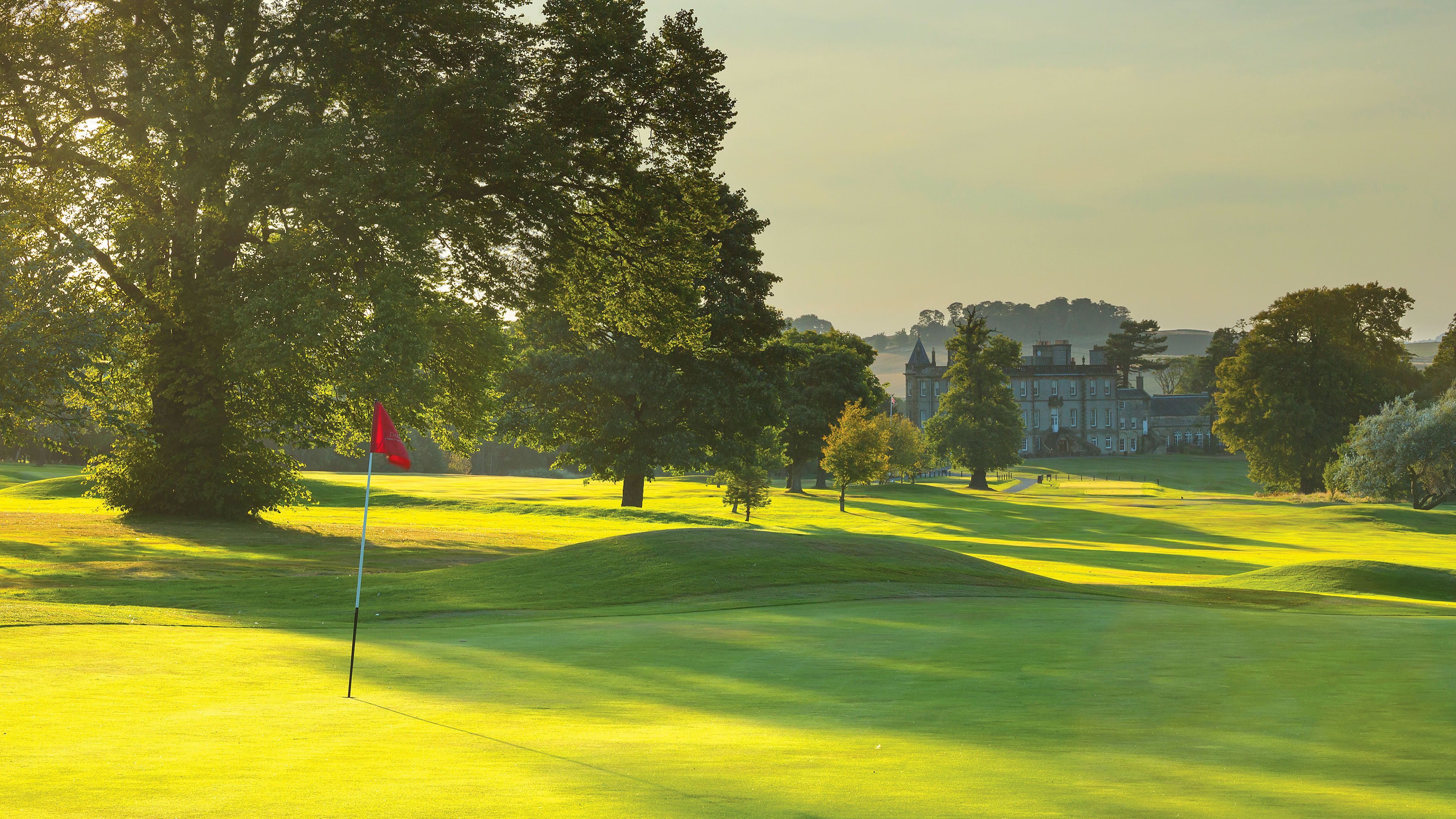 Un terrain de golf avec un mât de drapeau, des arbres et un grand bâtiment en arrière-plan.