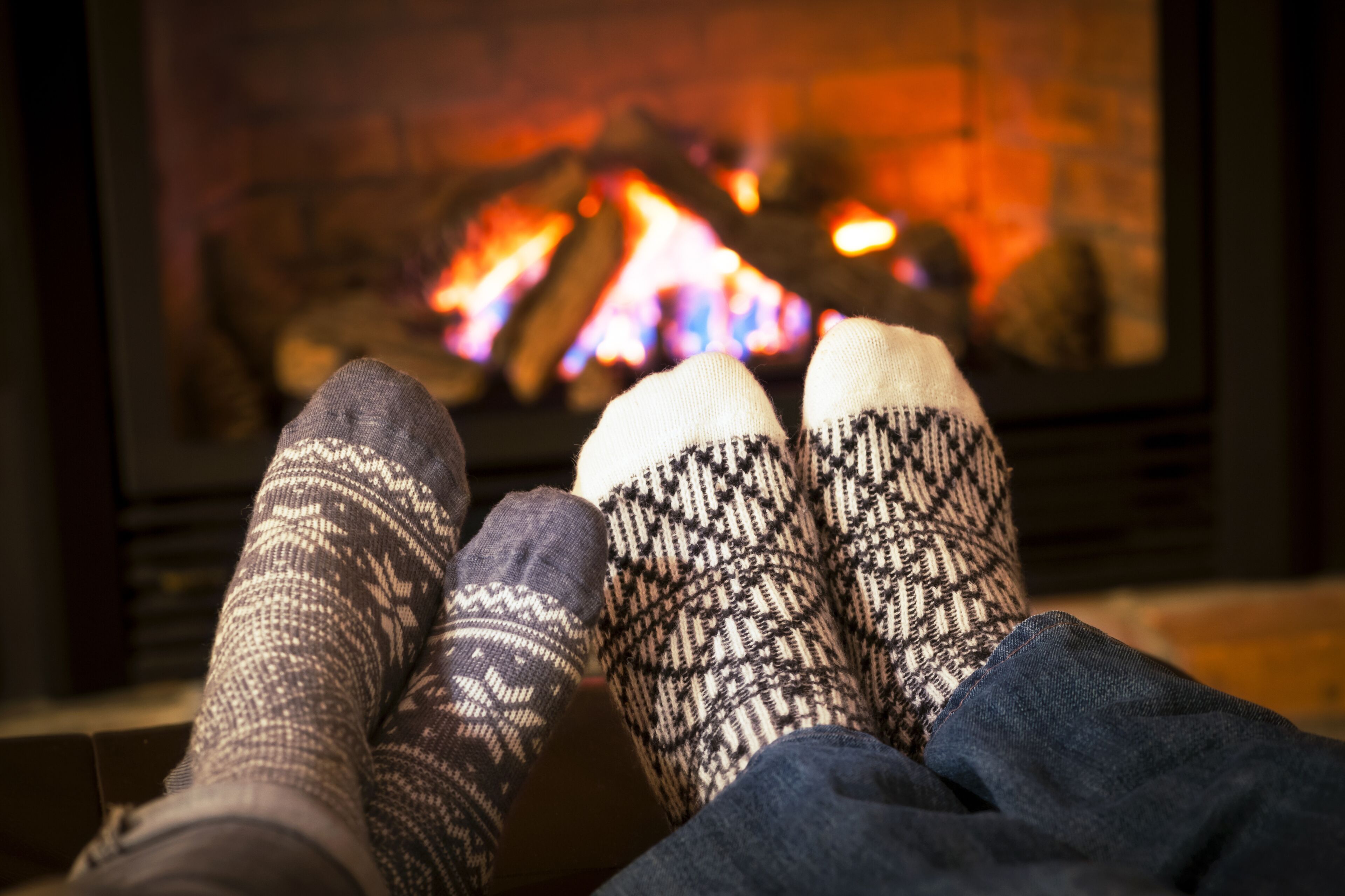 A fireplace with a fire, a person's legs with patterned socks, and a dark-colored sofa.