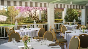 A covered outdoor dining area with tables set for a meal, surrounded by chairs, and overlooking a garden with blooming trees.