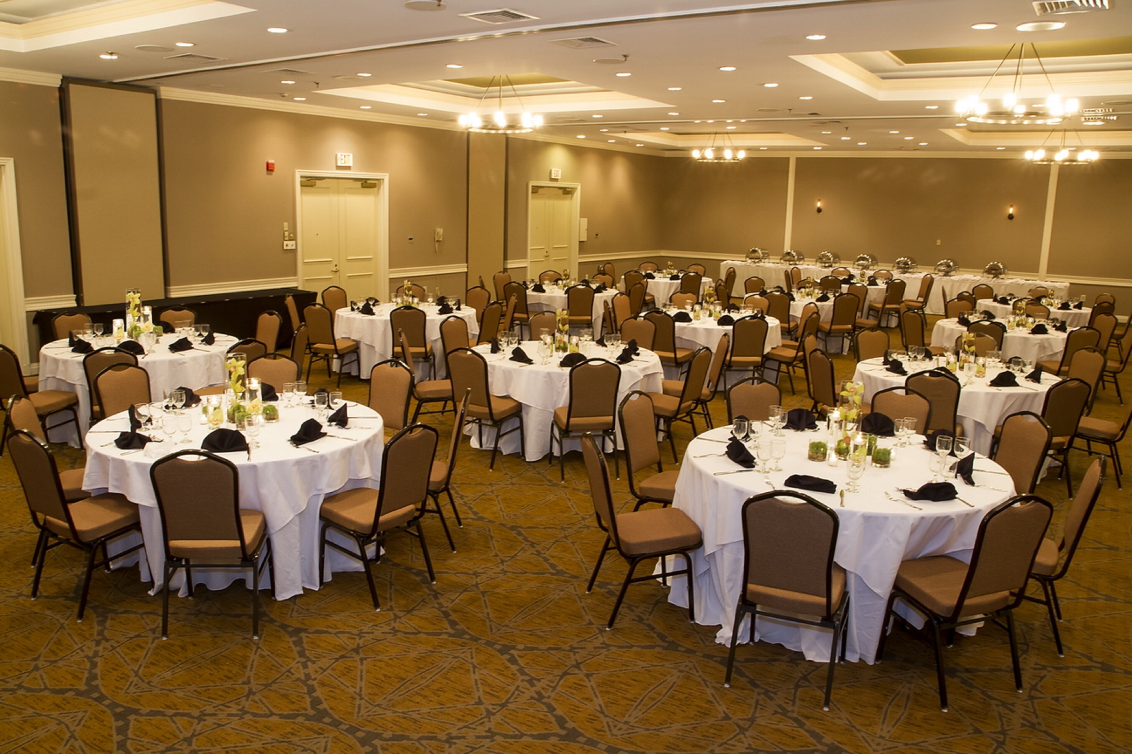 A large banquet hall with multiple round tables, each set with white tablecloths, chairs, and basic table settings.