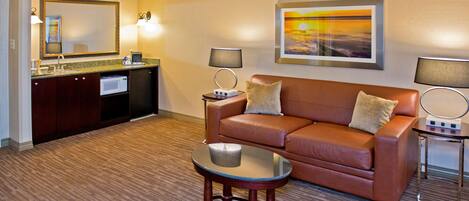 A hotel room with a brown leather sofa, a glass coffee table, a kitchen area with a microwave, and a painting on the wall.