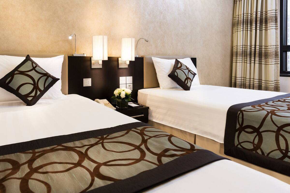 Ein Hotelzimmer mit zwei Betten, einem Nachttisch mit Lampe, einer Vase mit Blumen und einem Fenster mit Vorhängen.