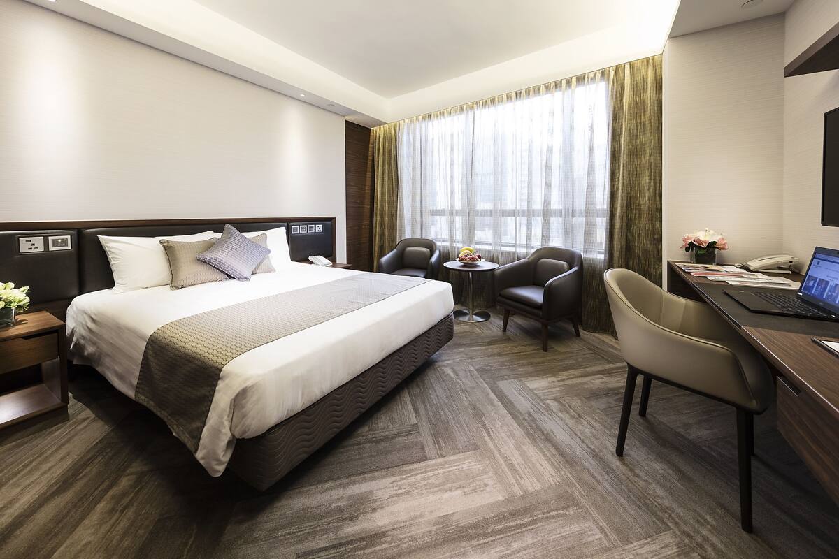 Ein Hotelzimmer mit einem großen Bett, zwei Sesseln, einem Schreibtisch mit einem Laptop und einem kleinen Tisch mit Blumen in einer Vase.