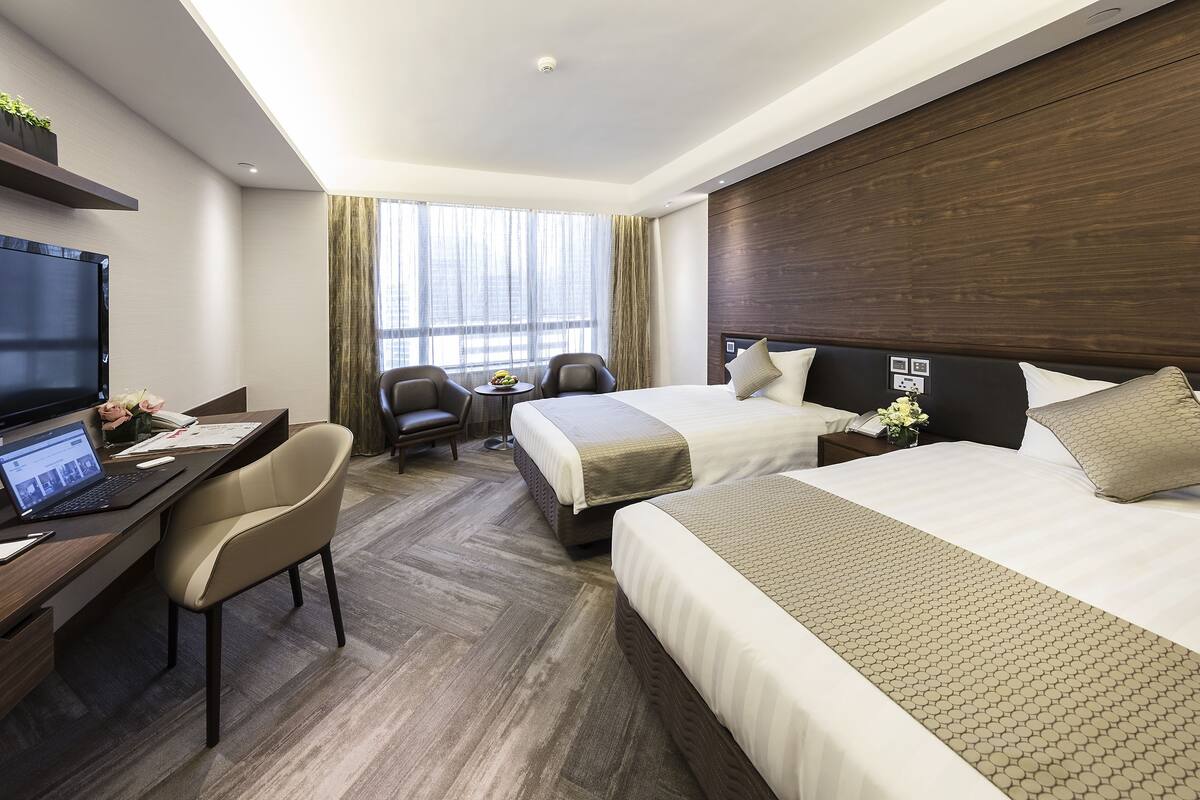 Ein Hotelzimmer mit zwei Betten, einem Schreibtisch, einem Laptop, einem Stuhl und einem Fernseher.