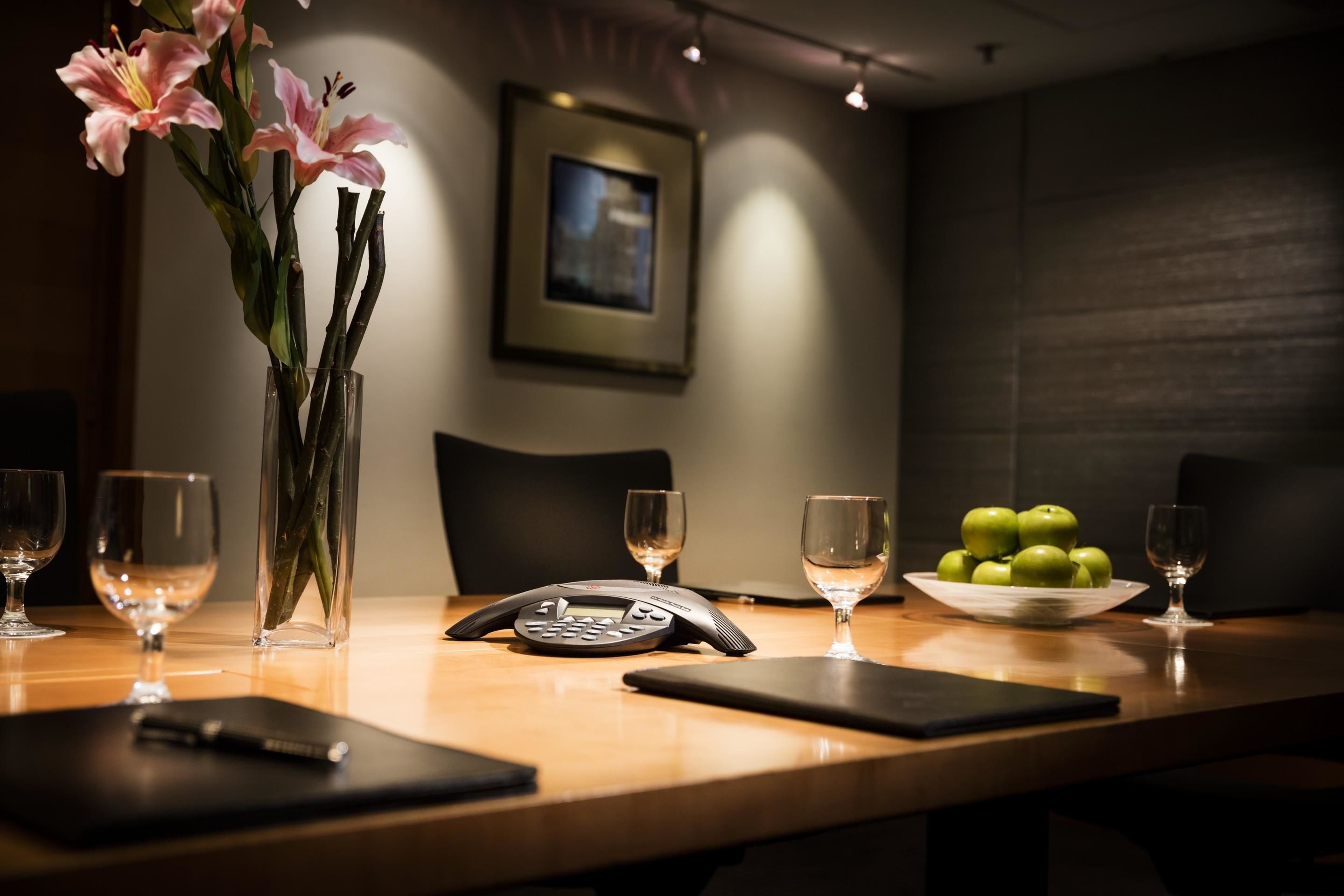 A conference table with a vase of flowers, a phone, a bowl of apples, and wine glasses.