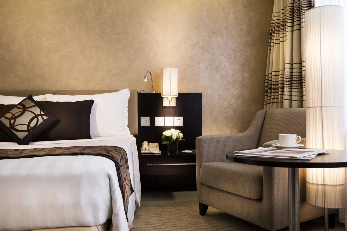 Ein Hotelzimmer mit einem Bett, einem Nachttisch mit Lampe, einem Sessel und einem kleinen Tisch mit Tasse und Zeitung.