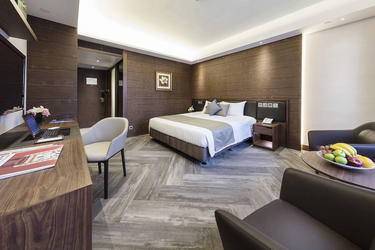 Ein modernes Hotelzimmer mit einem großen Bett, einem Schreibtisch mit einem Laptop, einem Stuhl und einem Esstisch mit Obst.