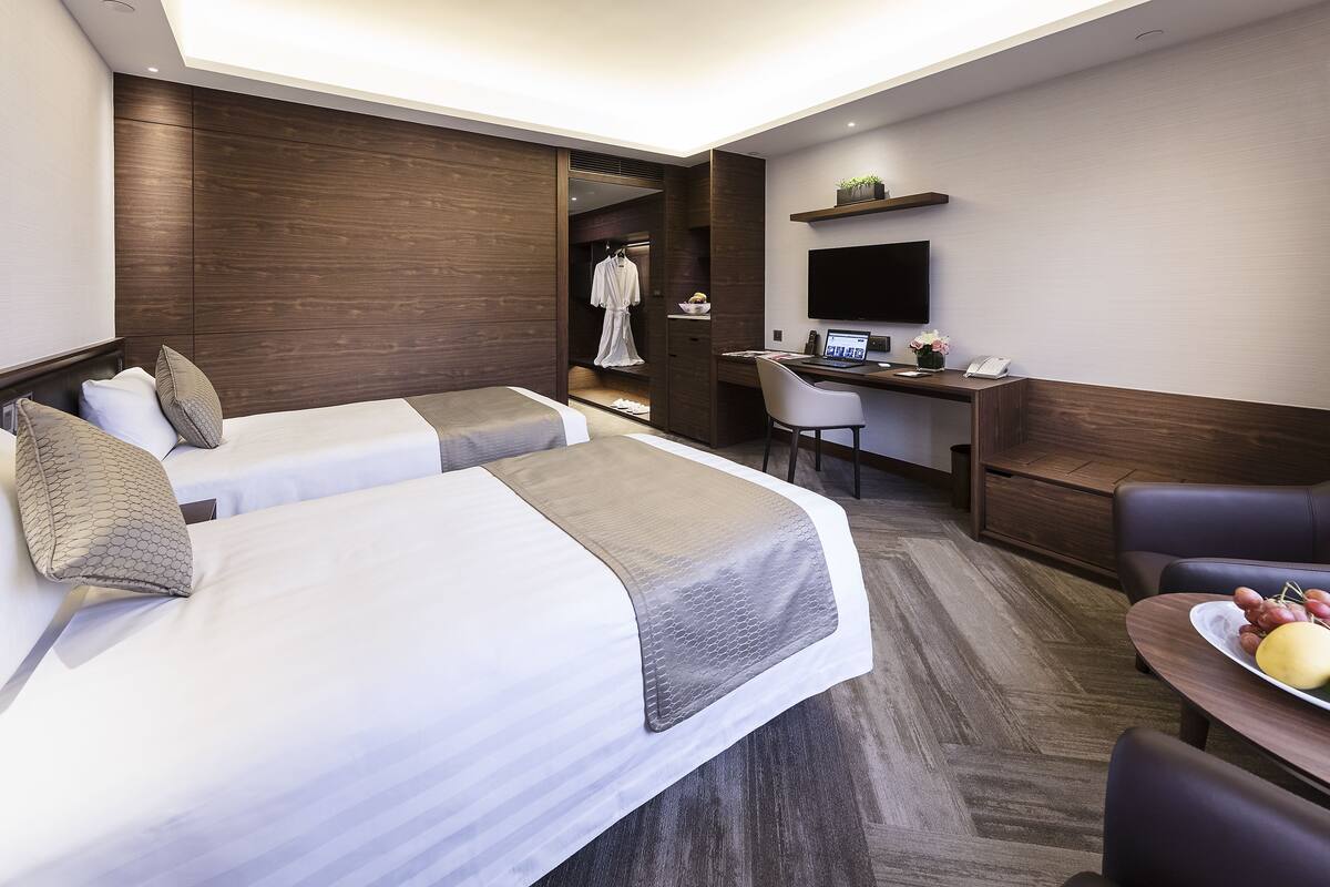Ein Hotelzimmer mit zwei Betten, einem Schreibtisch mit Computer, einem Fernseher und einem kleinen Tisch mit Obst.
