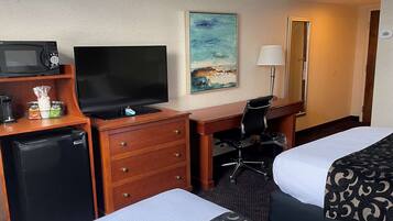 A hotel room with a desk, a television, a mini-fridge, a chair, two beds, a painting, a lamp, and a mirror.