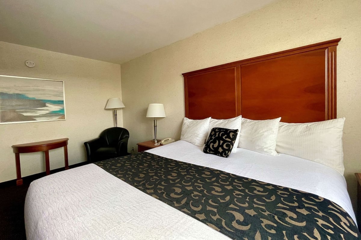 A hotel room with a large bed, two bedside tables, a chair, a painting on the wall, and a wooden headboard.