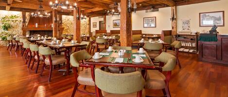 Une salle à manger spacieuse, avec des tables et des chaises en bois, des lustres et un mur en pierre.