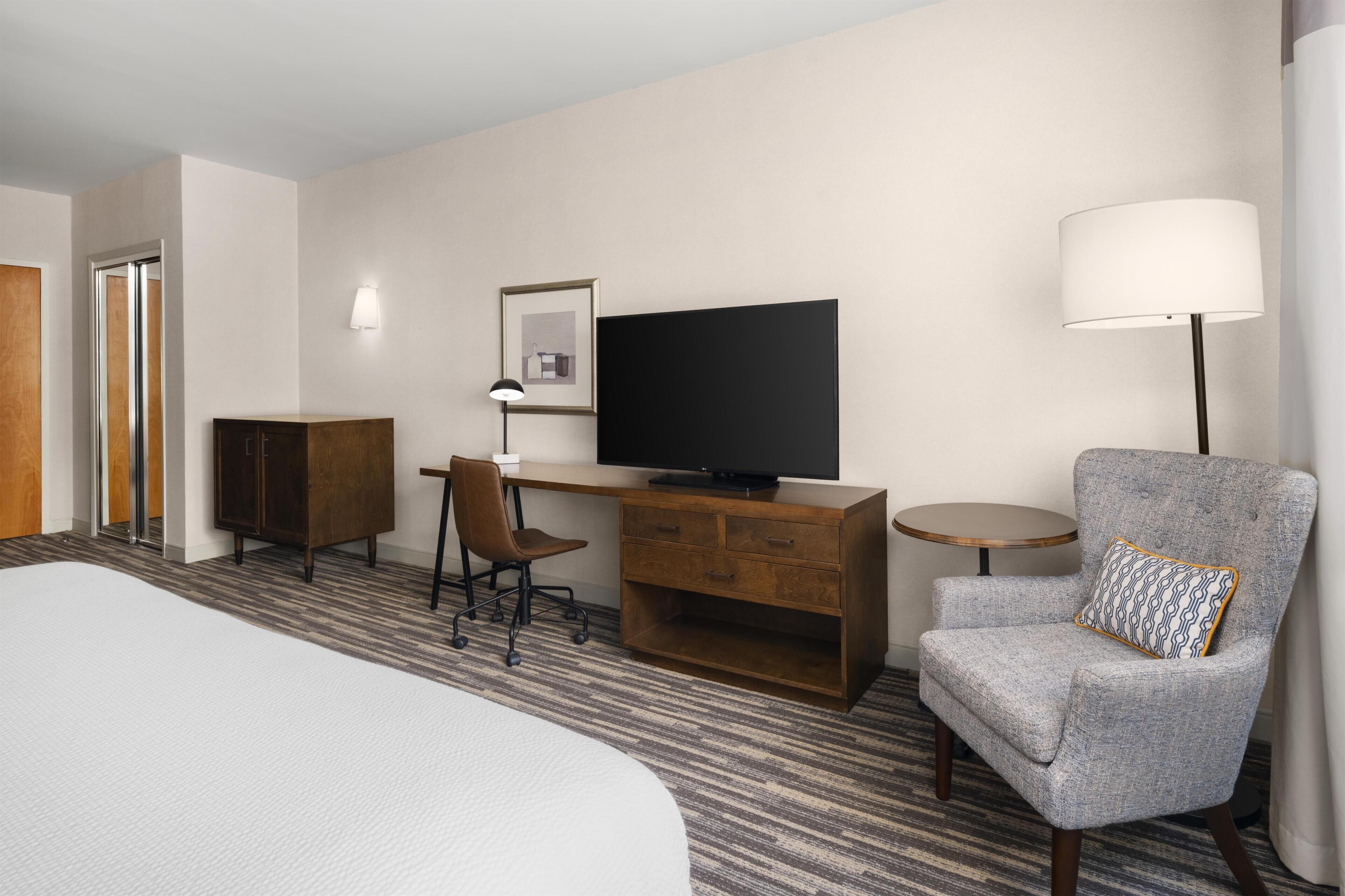 Habitación de hotel con cama, escritorio con televisor, silla, lámpara y armario.