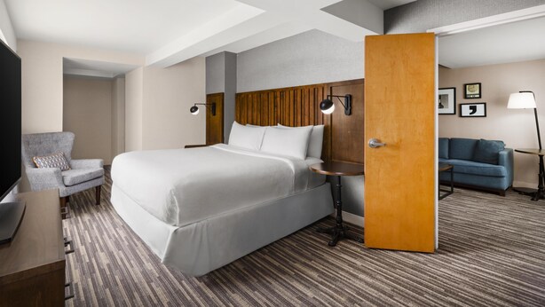 Habitación de hotel con una cama grande, zona de estar con una silla y sofá, escritorio y puerta que da a otra habitación.