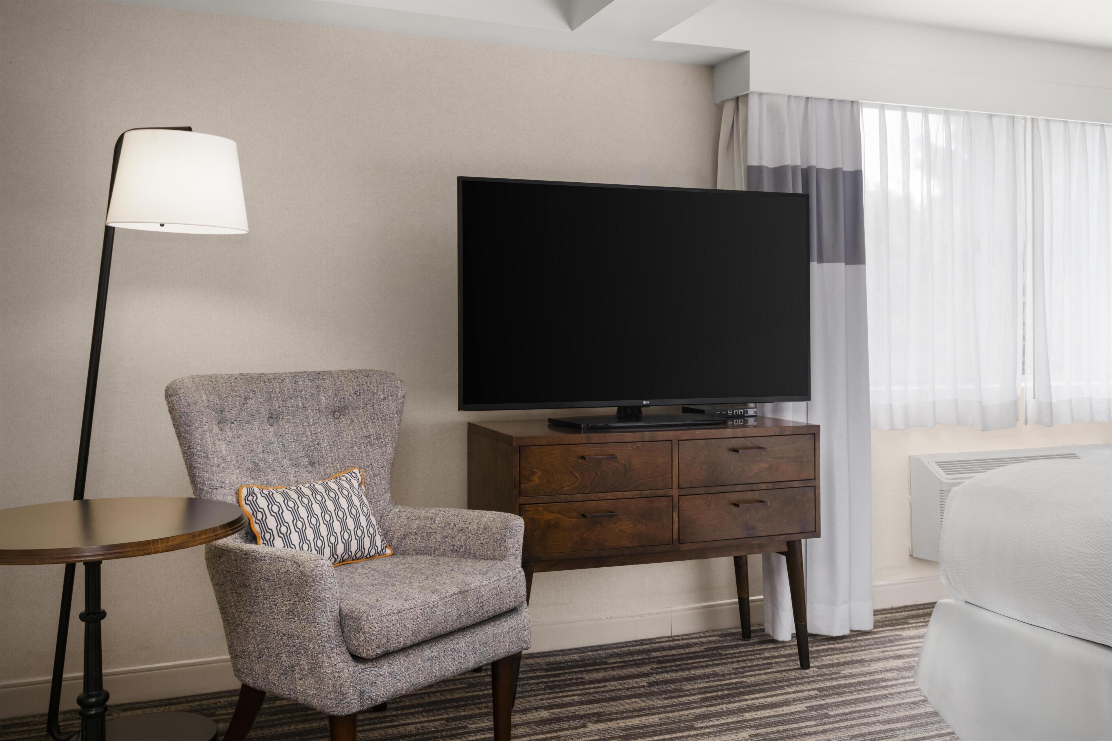 Een hotelkamer met een grijze fauteuil, een klein rond tafeltje, een flatscreen televisie op een houten kast en een bed met wit beddengoed.