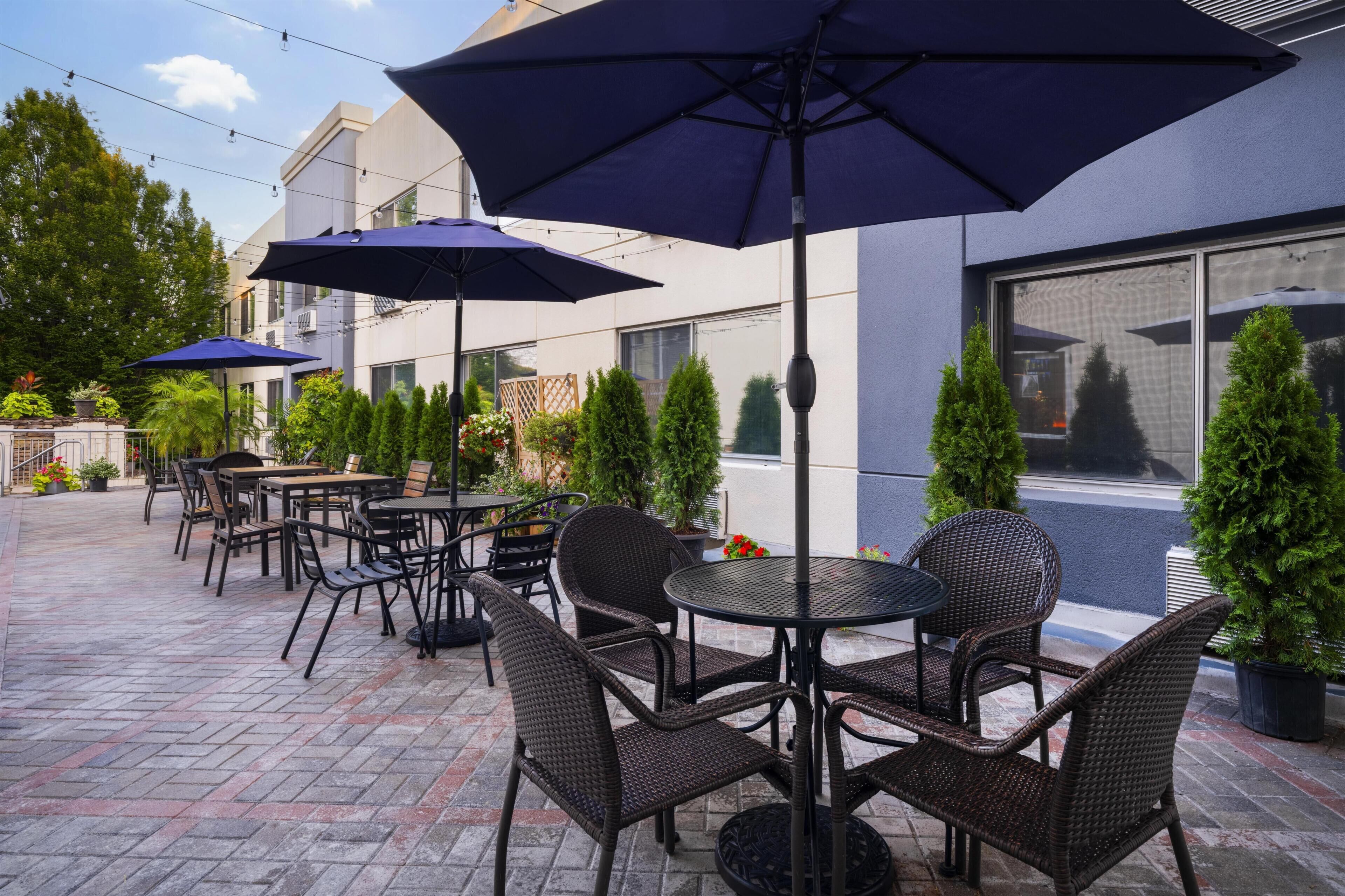Un espace extérieur aménagé en terrasse, avec plusieurs tables et chaises, dont certaines sont dotées de parasols, ainsi que des plantes en pot.