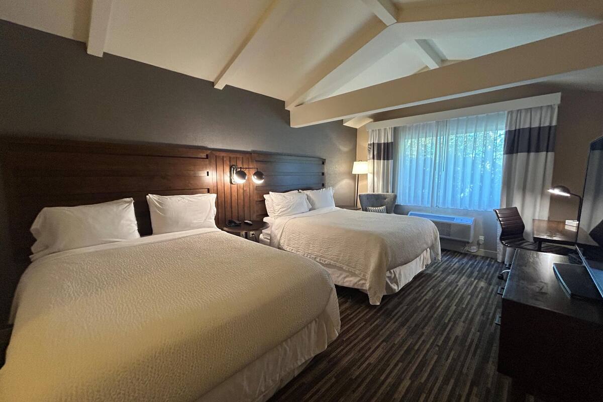 A hotel room with two beds, a desk, and a chair. There is a window with curtains, a lamp, and a wall-mounted light fixture.