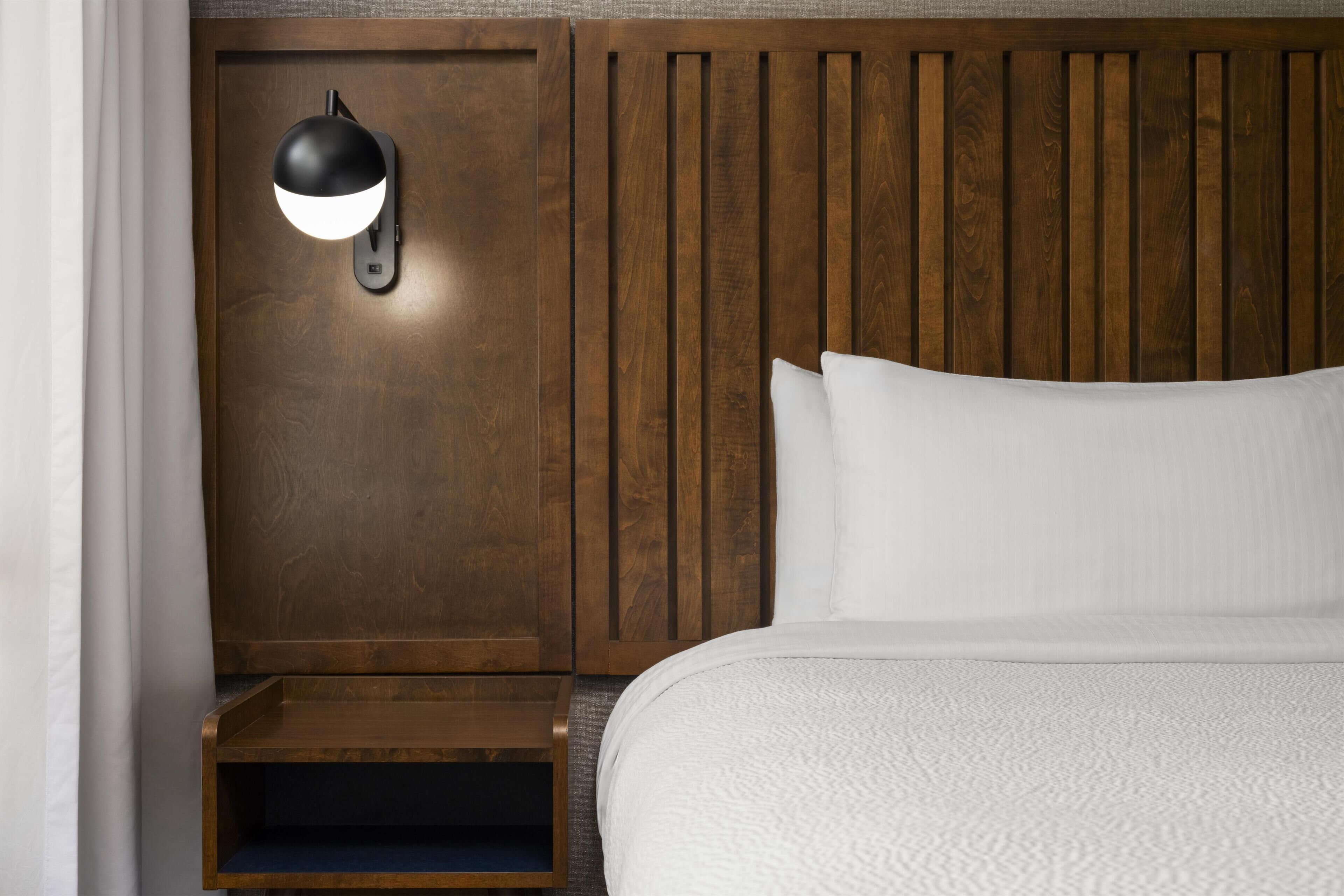 A hotel room with a wooden headboard, a bedside table, a lamp, and a bed with white bedding.