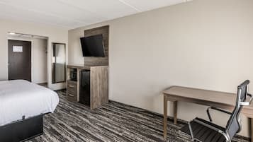 A hotel room with a bed, a desk, a chair, a television mounted on a wooden panel, and a carpeted floor.