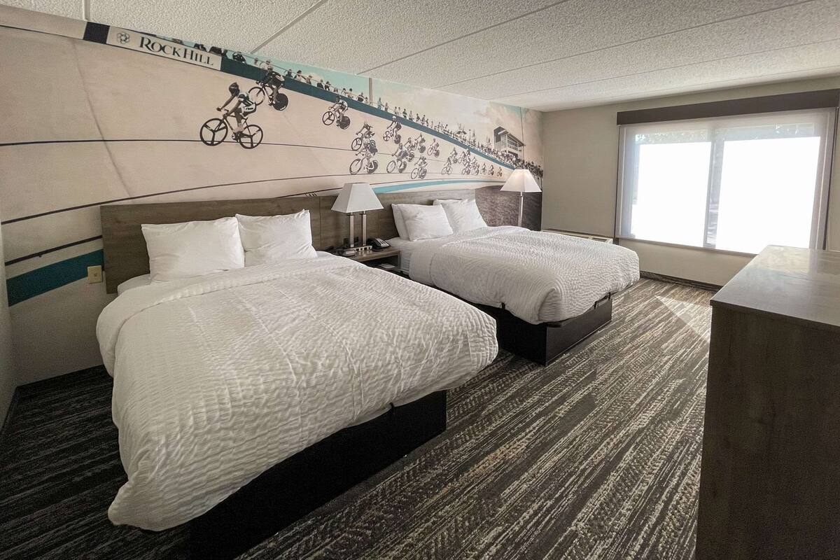 A hotel room with two beds, a large window, and a wall mural of cyclists.