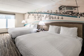 Habitación de hotel con dos camas, un ventanal grande, un mural en la pared de una carrera ciclista y una lámpara.
