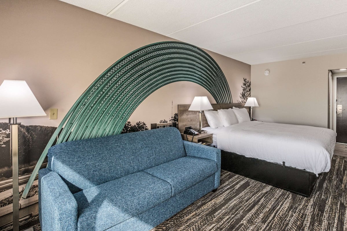 A hotel room with a blue sofa, a bed with white bedding, a curved green wall feature, and a patterned carpet.