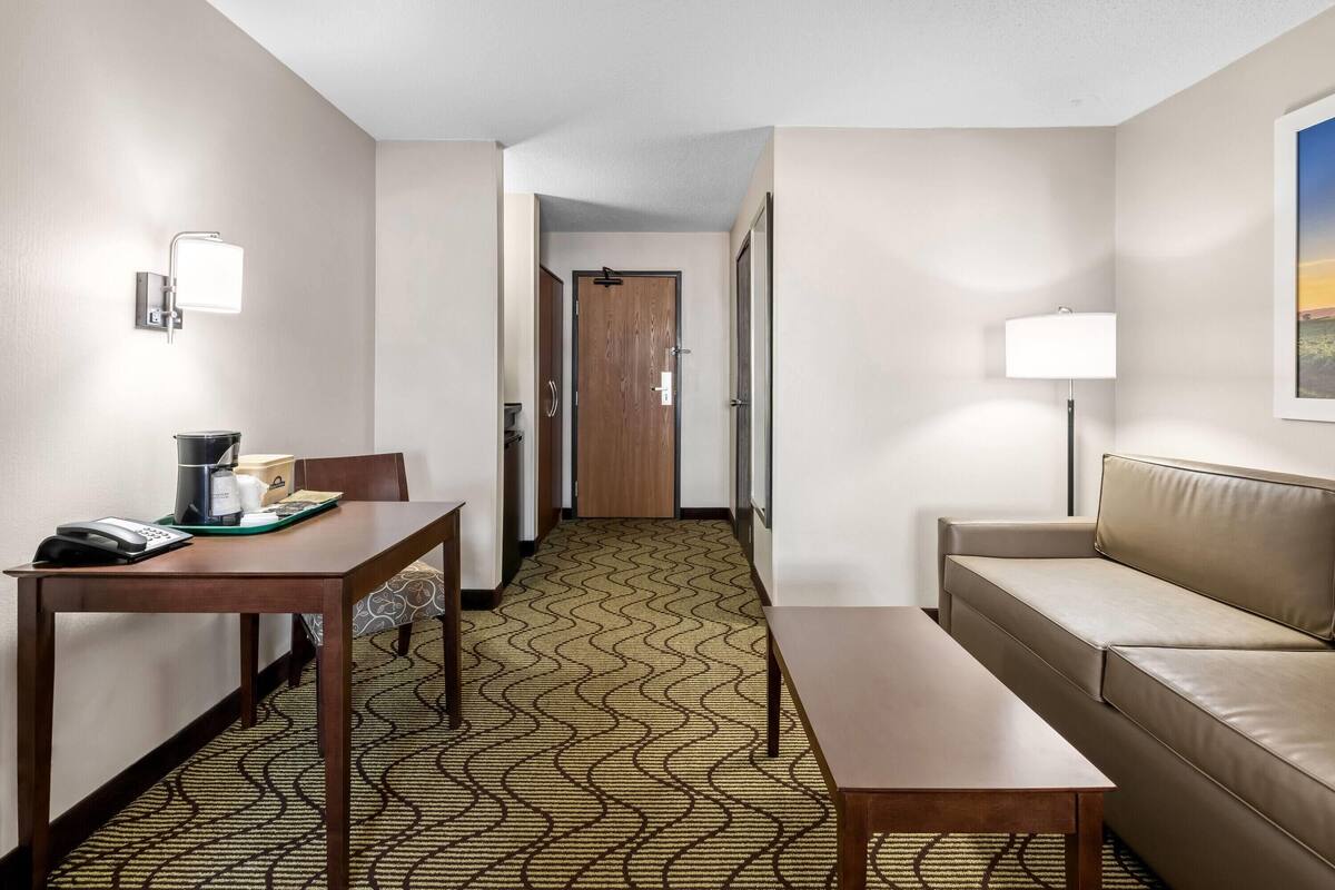 Habitación de hotel con un escritorio de madera, un sofá, una alfombra estampada y una puerta que da a otra habitación.