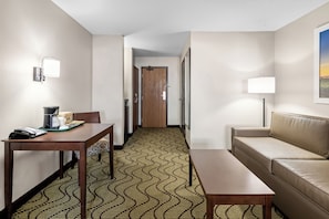 Una habitación de hotel con un escritorio de madera, un sofá, una alfombra estampada, una puerta y una lámpara.