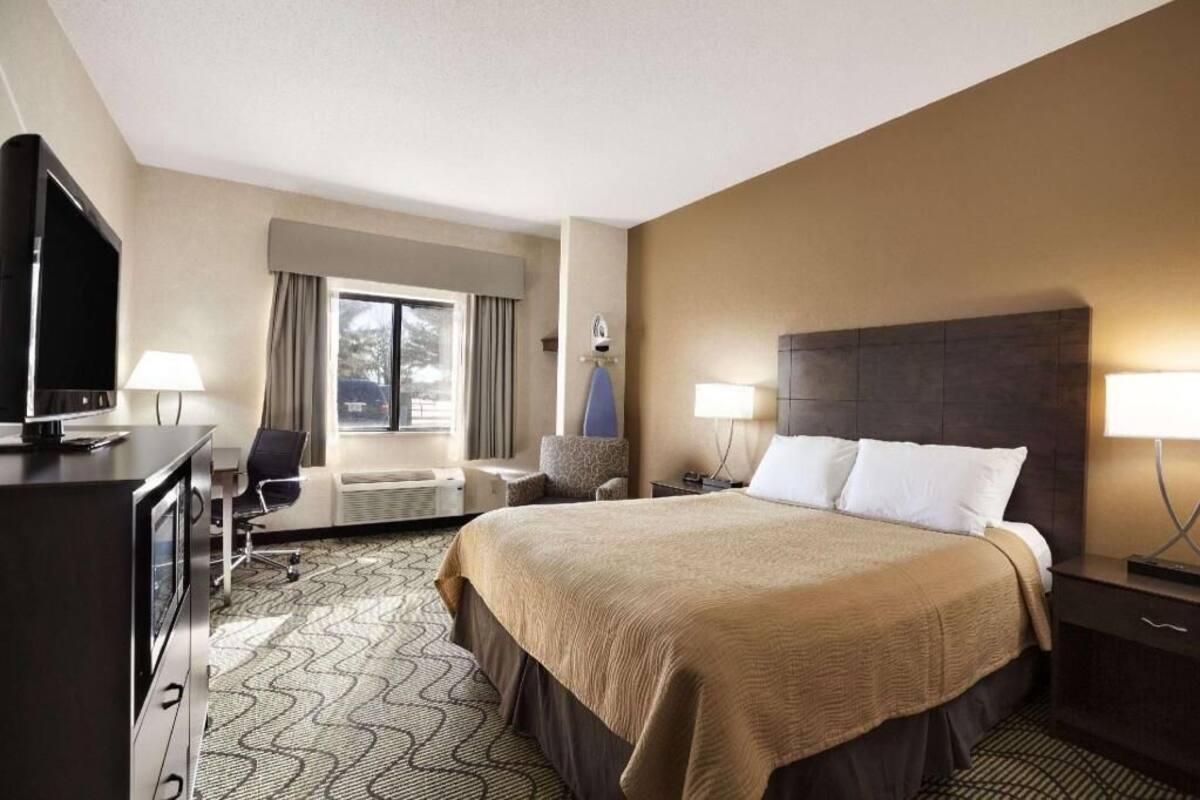 Habitación de hotel con una cama grande, un escritorio con televisor, una silla, una ventana con cortinas y una lámpara.