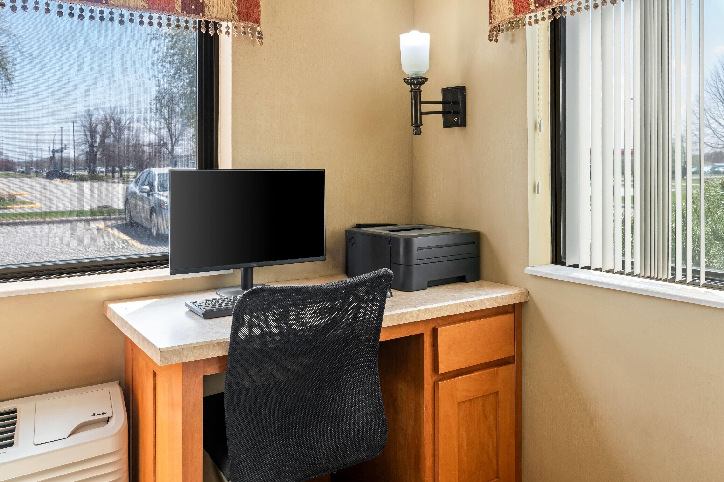 Ein Schreibtisch mit Computerbildschirm, Tastatur und Drucker, ein Stuhl und ein Fenster mit Jalousien.