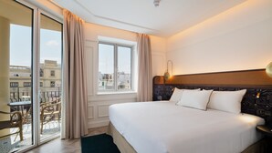 Habitación de hotel con una cama grande, vistas de la ciudad a través del ventanal y un balcón con mobiliario exterior.