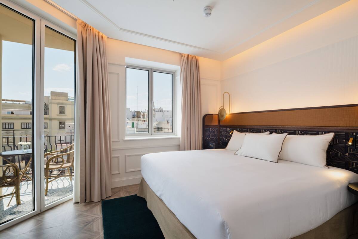 A hotel room with a large bed, a view of the city through the window, and a balcony with outdoor furniture.