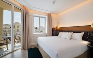 Quarto de hotel com uma cama grande, vista da cidade pela janela e varanda com mobiliário exterior.