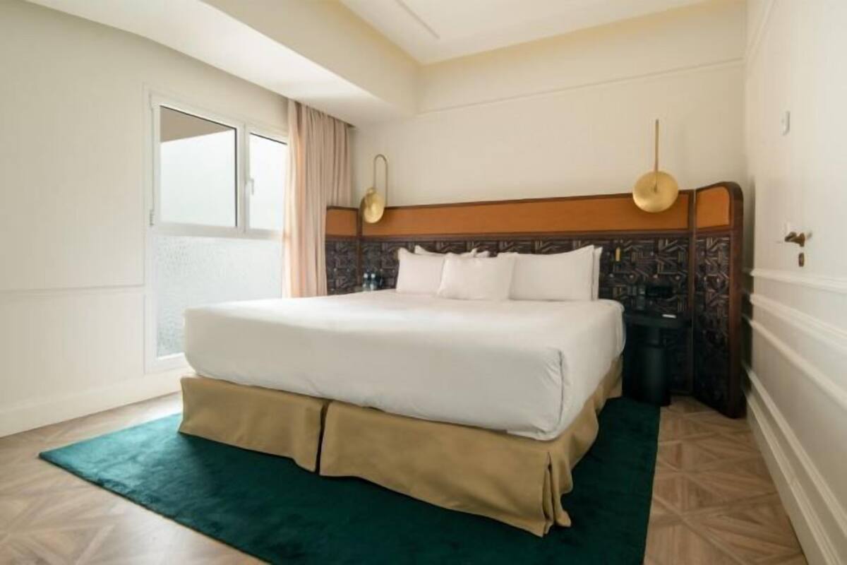 A hotel room with a large bed, a bedside table, a nightstand, a window with curtains, and a wall-mounted light fixture.