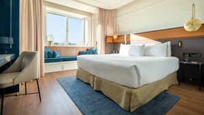 Habitación de hotel con una cama grande, un escritorio, una silla, un ventanal con vistas y una mesita de noche con lámpara.