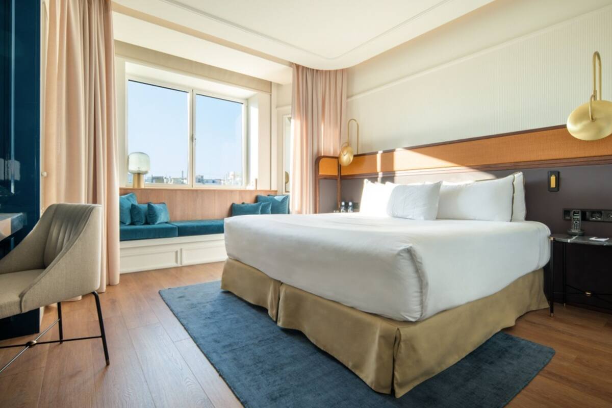 A hotel room with a large bed, a desk, a chair, a window with a view, and a bedside table with a lamp.
