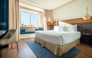 Quarto de hotel com uma cama grande, uma escrivaninha, uma cadeira, uma janela com vista e uma mesinha de cabeceira com abajur.