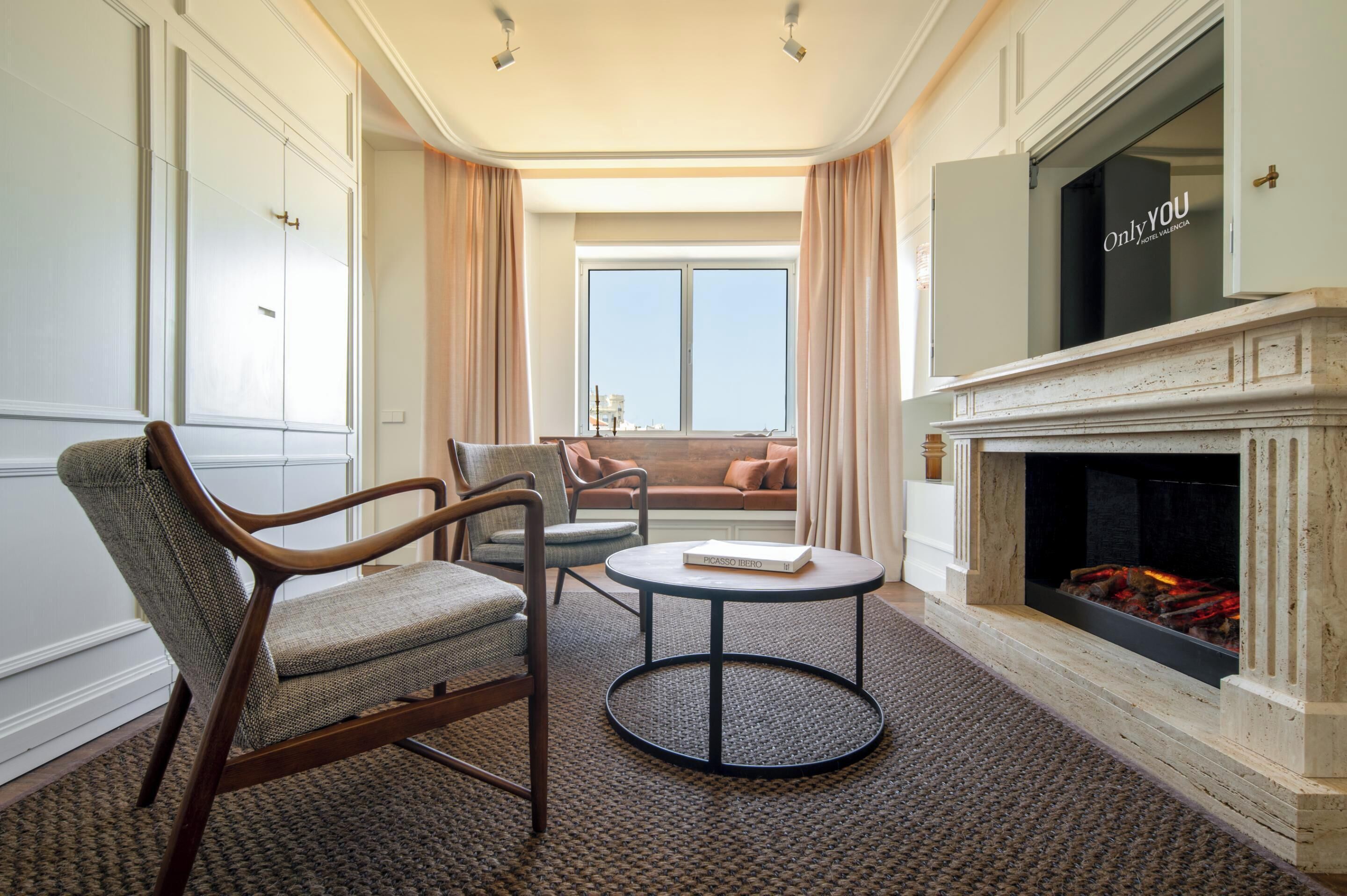 A hotel room with a fireplace, a sofa, two armchairs, a round table, and a large TV screen.