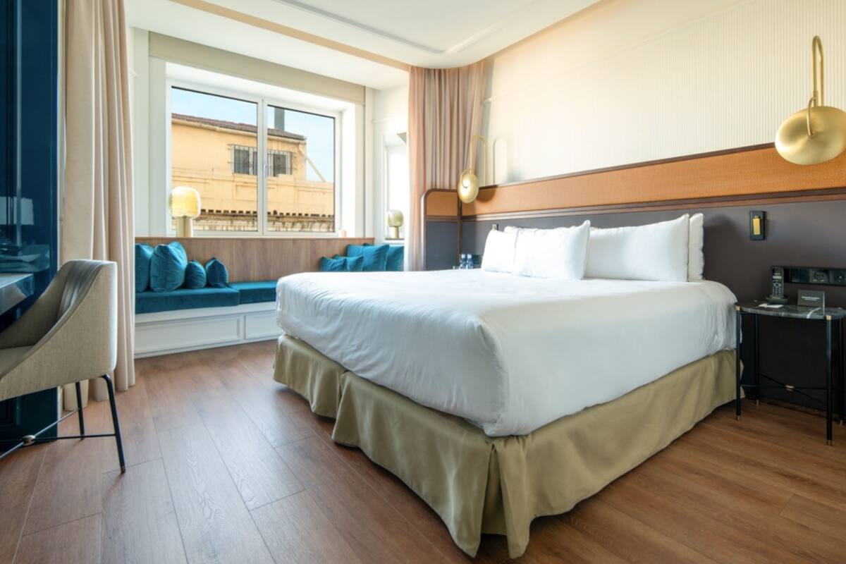 A hotel room with a large bed, a desk with a chair, a window with a view of buildings, and a bedside table with a lamp.